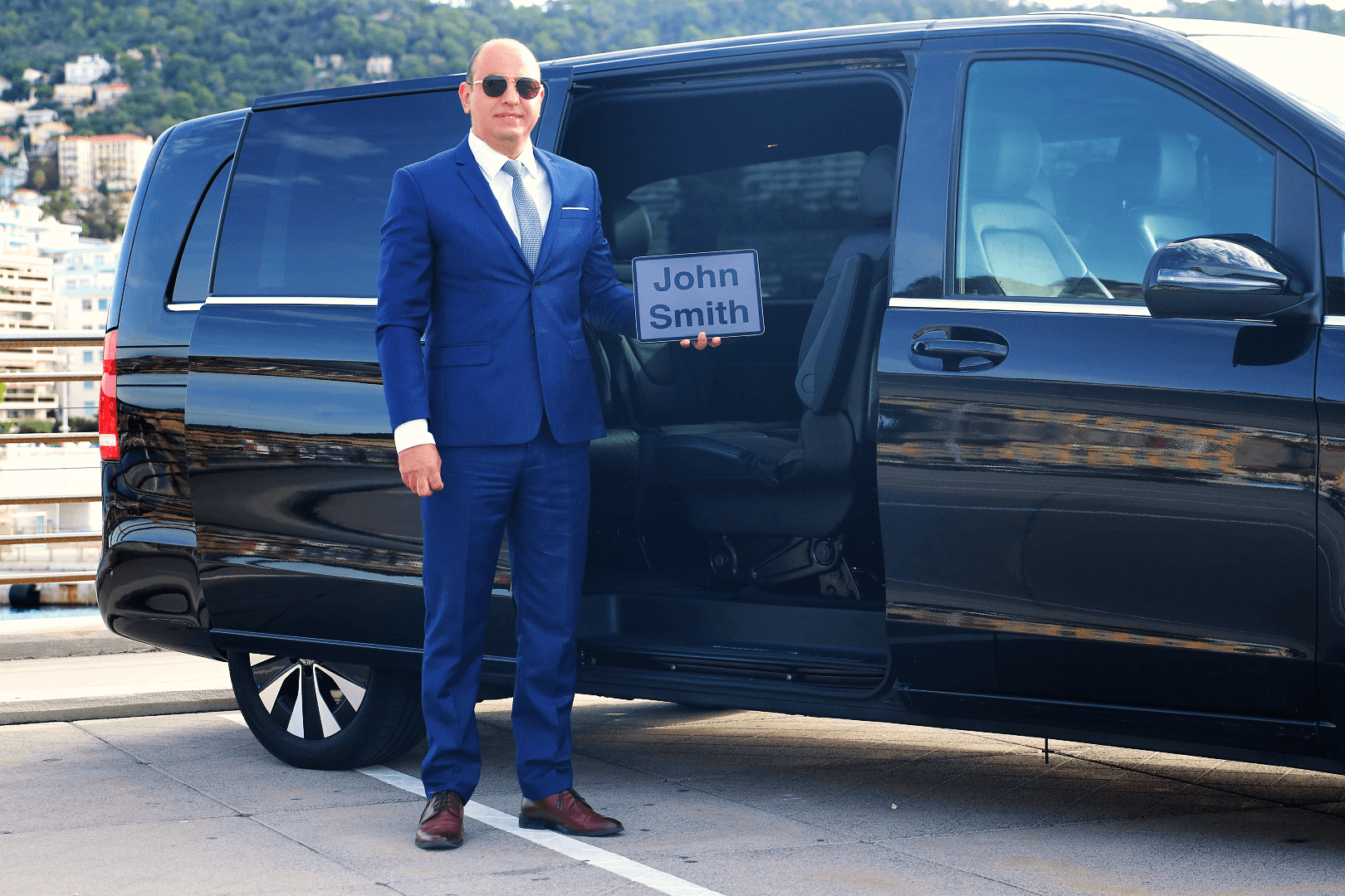 Professional chauffeur in a blue suit and sunglasses standing by an open black van door, holding a “John Smith” name sign